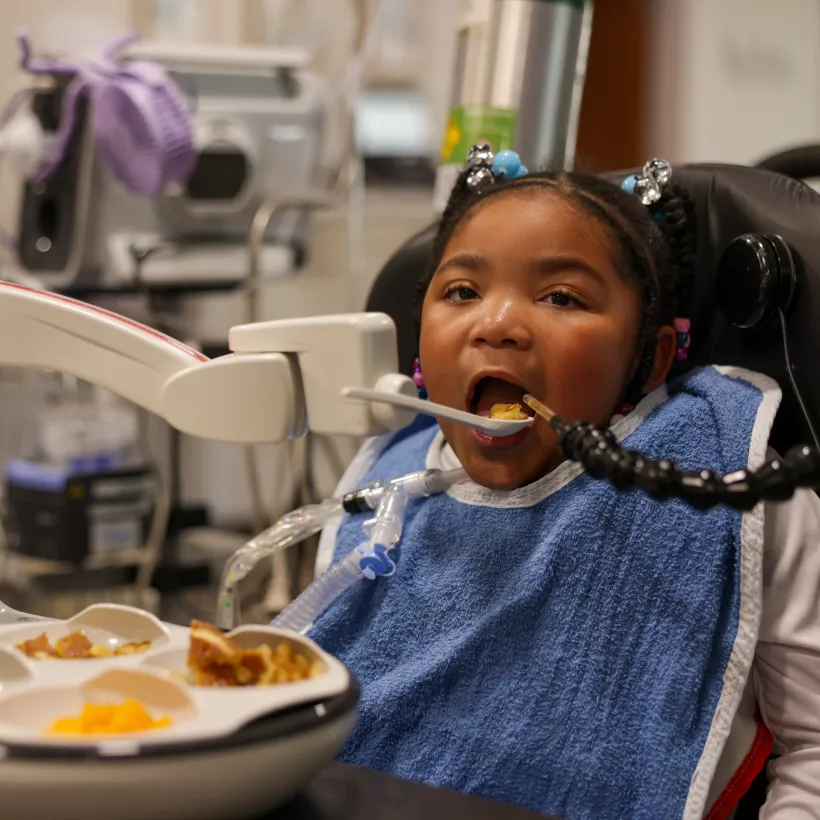 A young girl sits in a supportive wheelchair wearing a blue bib, connected to a ventilator through a tracheostomy tube.