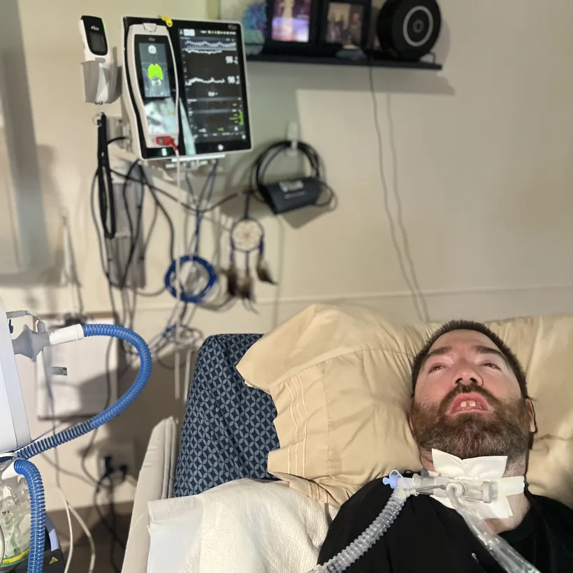 A man with a tracheostomy connected to a ventilator rests in a medical chair beside monitoring equipment and respiratory tubing, with family photos displayed on shelves behind him.